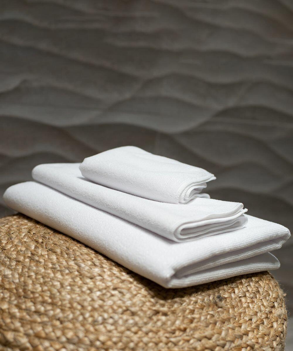 A stack of Towel City TC018 white folded towels placed on a woven wicker stool with a textured wall in the background.
