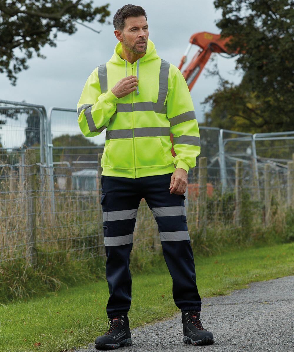A man in a neon yellow Yoko YK012 high-visibility hoodie and work pants stands outdoors near a metal fence and construction equipment.