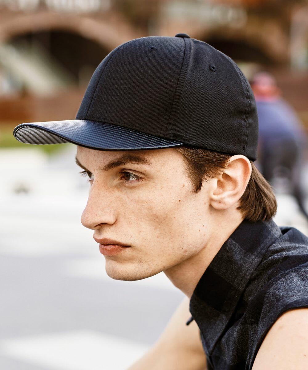 A man with light skin and brown hair is seen in a profile view wearing a black and blue Flexfit By Yupoong YP079 cap. He is outdoors, sitting on a bench, with blurred buildings and people in the background.
