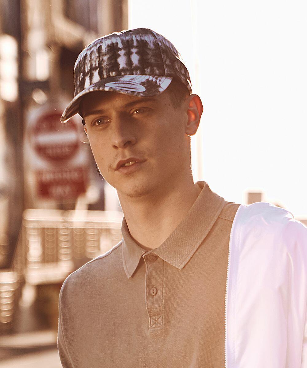 A young man wearing a Flexfit By Yupoong YP099 and a polo shirt stands outdoors with blurred street signs in the background.