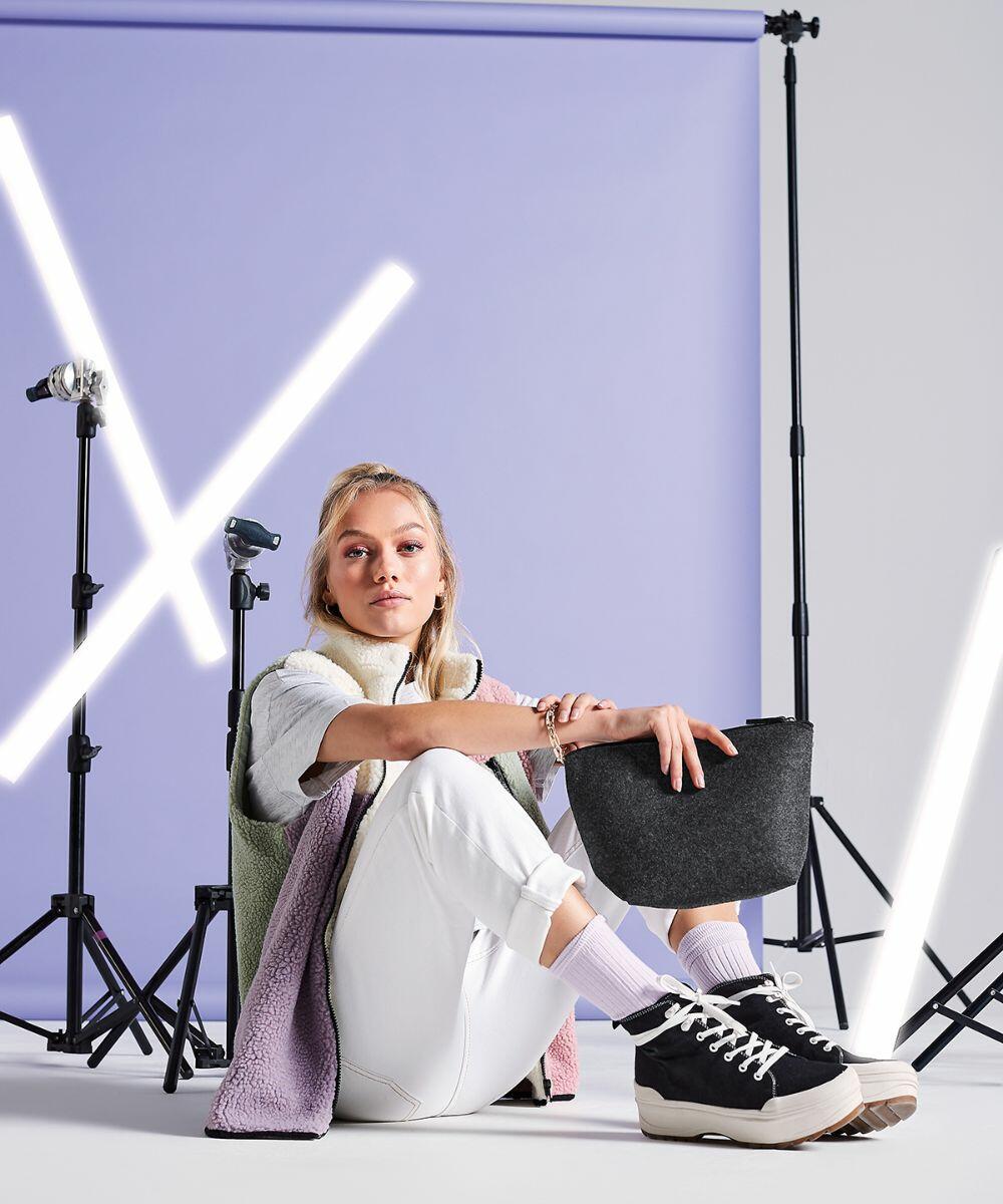 A person with blonde hair sits on the floor against a purple backdrop, holding a black Bagbase BG724. They wear a white outfit with black and white high-top sneakers. Studio lights on stands are arranged behind, with bright tubes illuminating the scene.