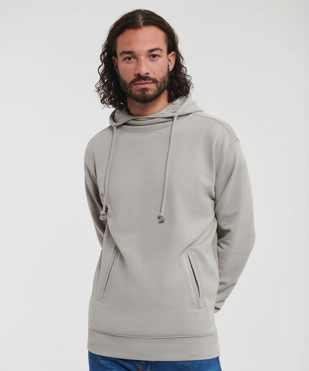 A man with long curly hair and a beard is wearing a Russell J209M light gray hoodie with front pockets and blue jeans. He stands against a plain white background.