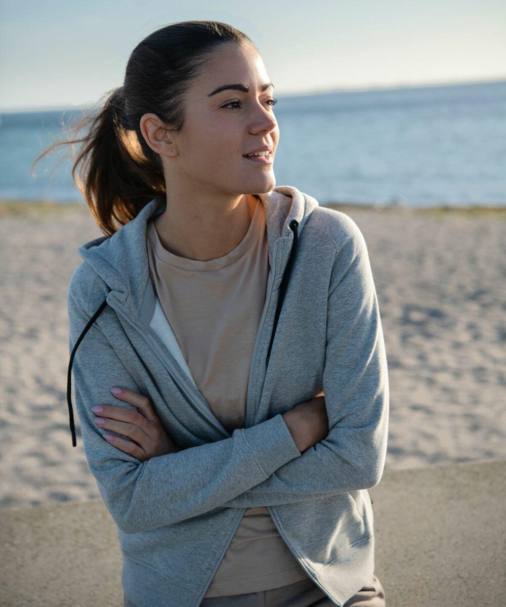 A woman with a ponytail wearing a Nimbus NP04F stands with her arms crossed, looking to the side, with a beach and ocean in the background.