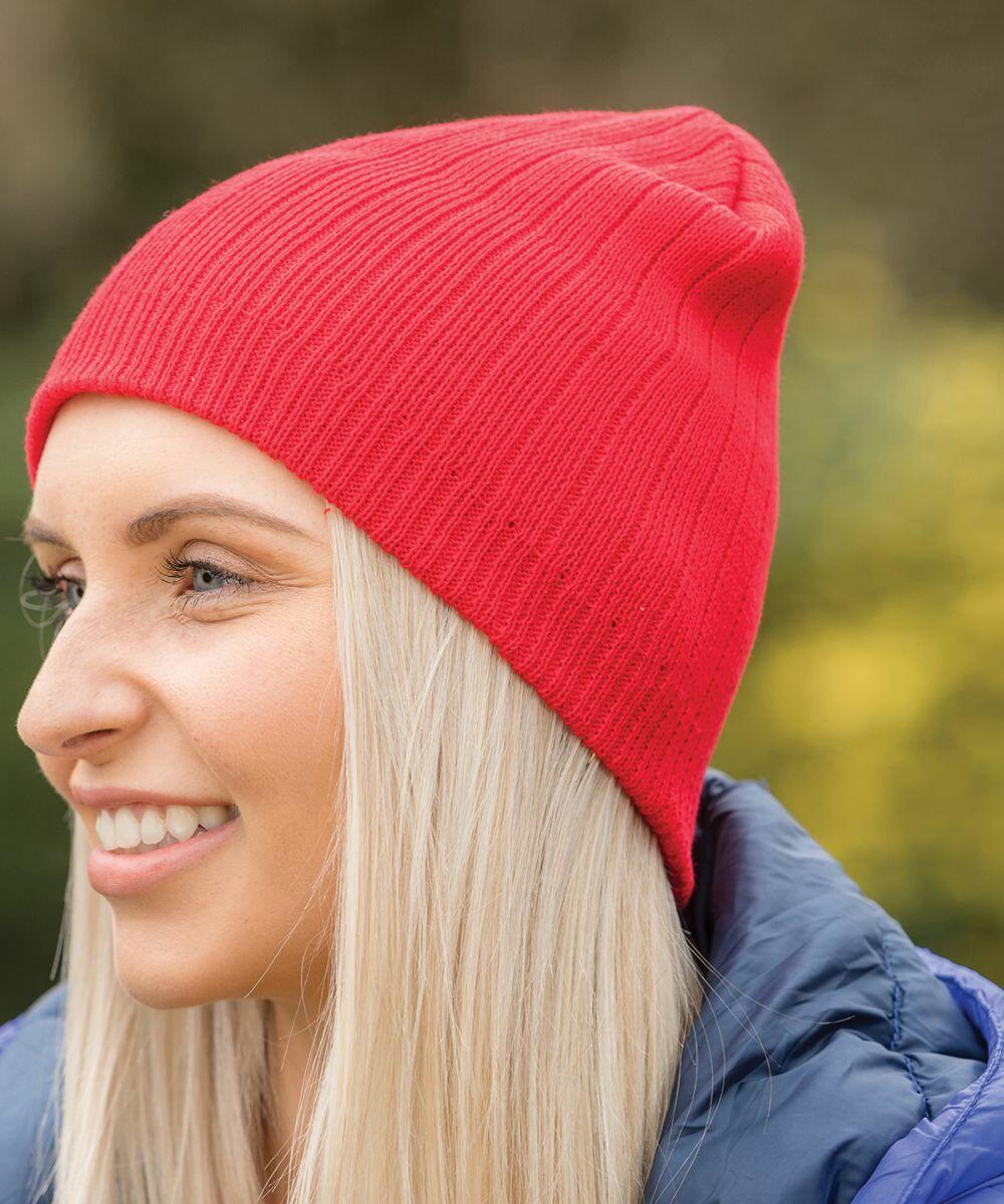 Person with long blonde hair wearing a bright red beanie and a blue RC074 jacket from Result, smiling while looking away from the camera.