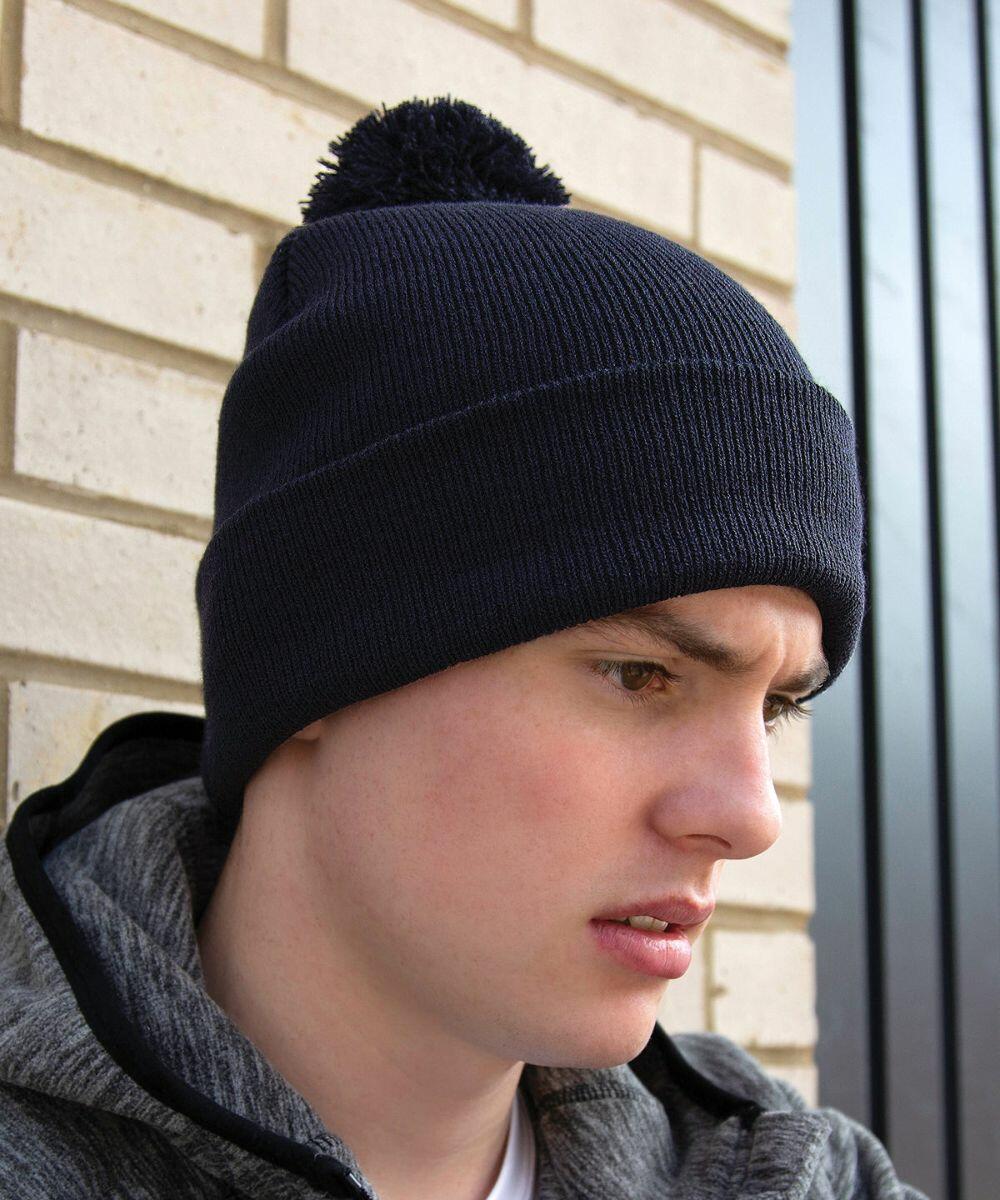 A person wearing a dark knitted beanie with a pompom and a grey RC28X jacket from Result stands against a brick wall, looking downward.