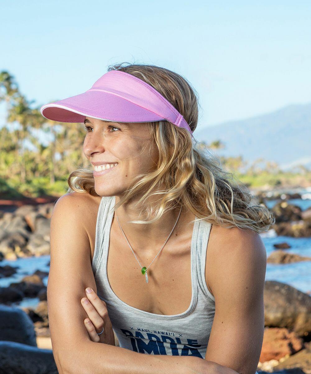 A woman with wavy blonde hair wearing a pink Result RC48X visor and a gray tank top smiles while standing on a rocky beach with trees and mountains in the background.