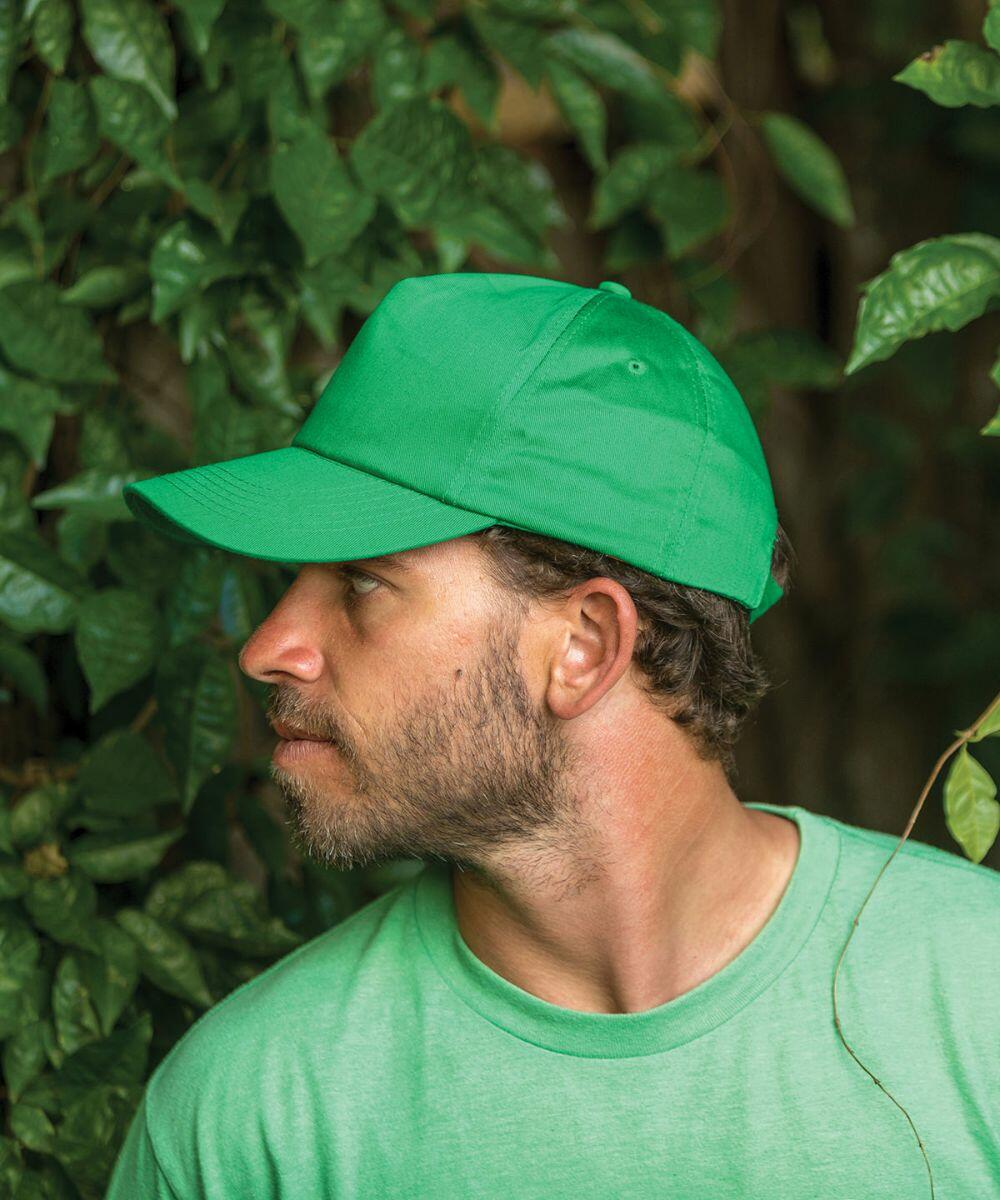 A man wearing a green cap and green shirt from Result's RC84X collection is looking to his left, standing against a backdrop of dense green foliage.
