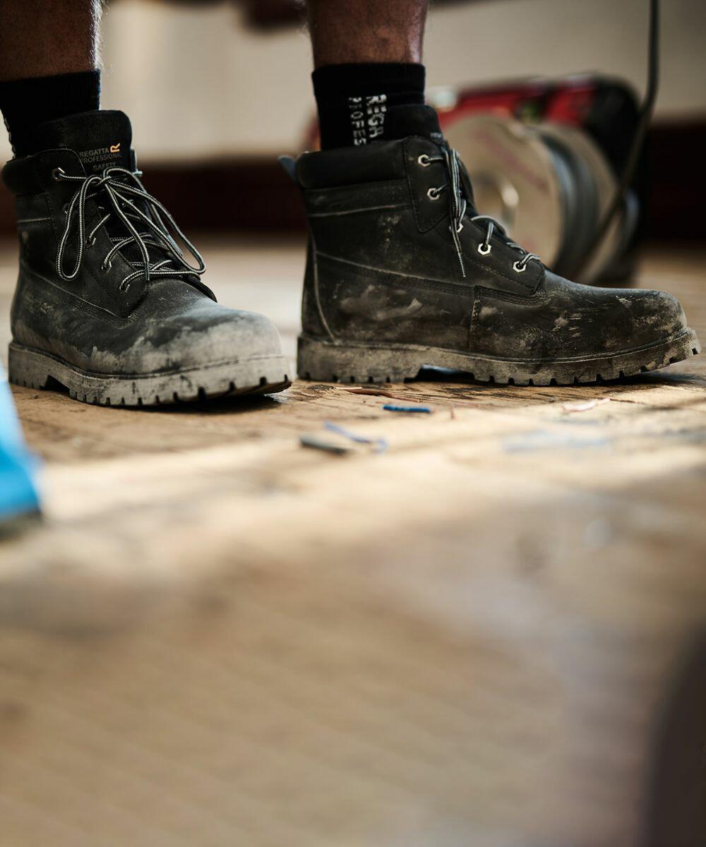 Close-up of a person wearing Regatta RG571 black work boots with mud on them, standing on a wooden floor with scattered nails.