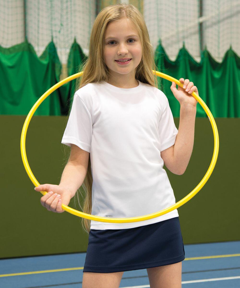 A young girl with long blonde hair stands inside a yellow hula hoop, wearing a white shirt and dark blue skirt, in a gymnasium with green curtains and a blue floor, holding the Spiro S261J.
