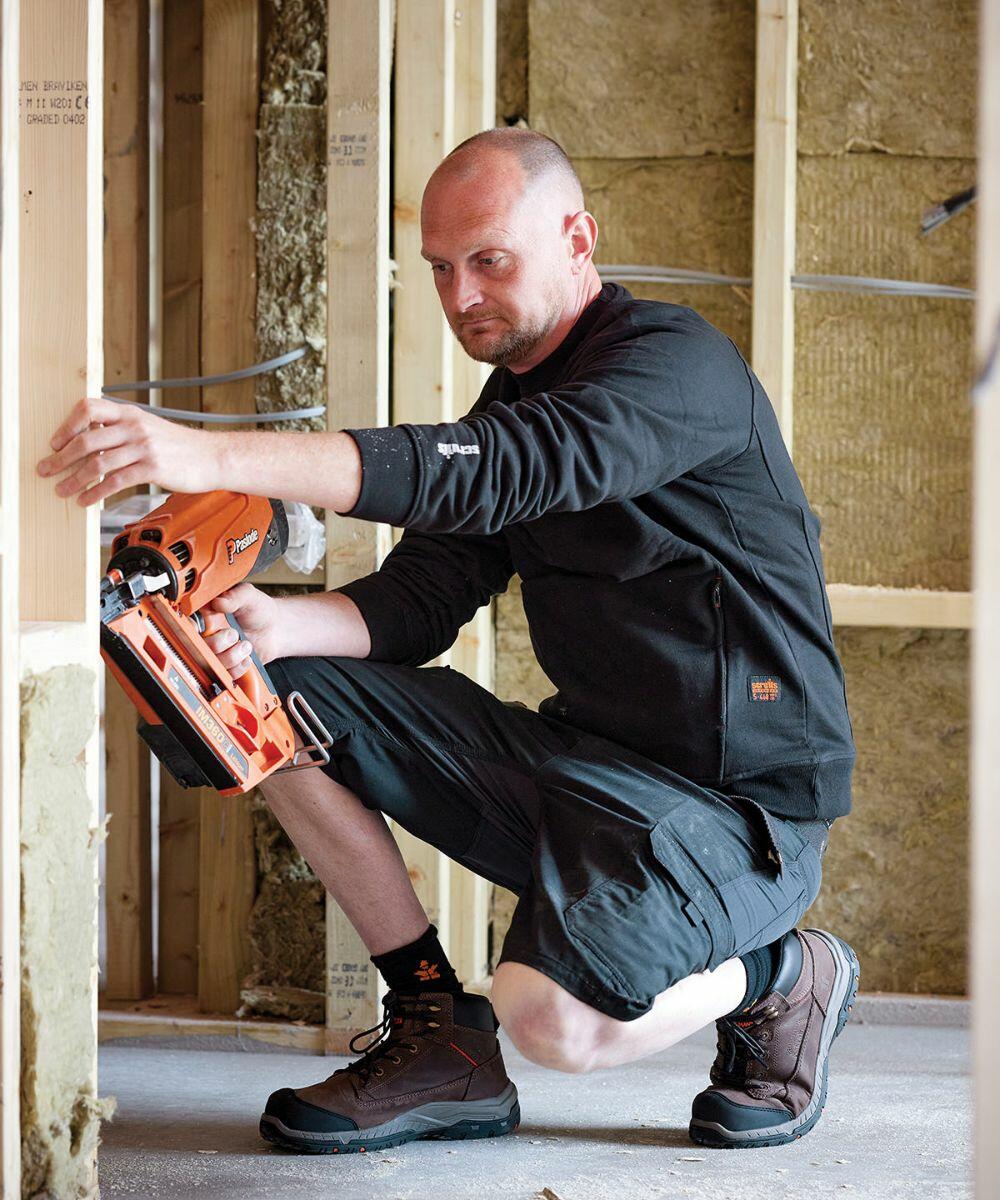 A man uses a SH002 by Scruffs while working on a construction project. He is wearing a black sweatshirt, black shorts, and work boots.