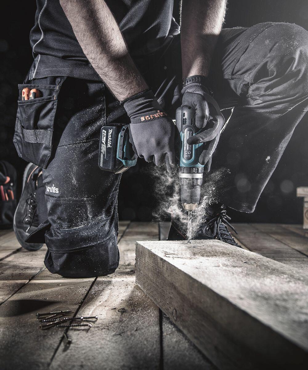 Person kneeling on a wooden floor, wearing work gloves and using a Scruffs SH027 cordless power drill on a piece of wood, with screws and tools visible nearby.