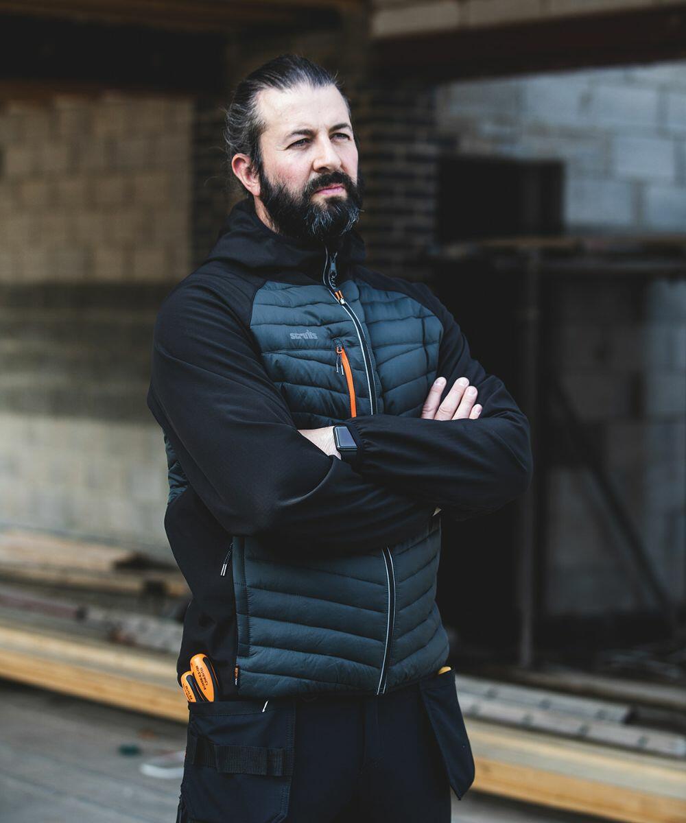 A person with a beard stands with arms crossed, wearing a black Scruffs SH033 jacket and black pants, in an industrial setting.