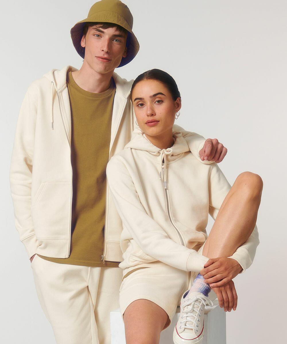 Two individuals dressed in cream-colored casual outfits. The person on the left wears a Stanley/Stella SX012 hat and stands with a neutral expression, while the person on the right sits with one leg on a block and smiles slightly.