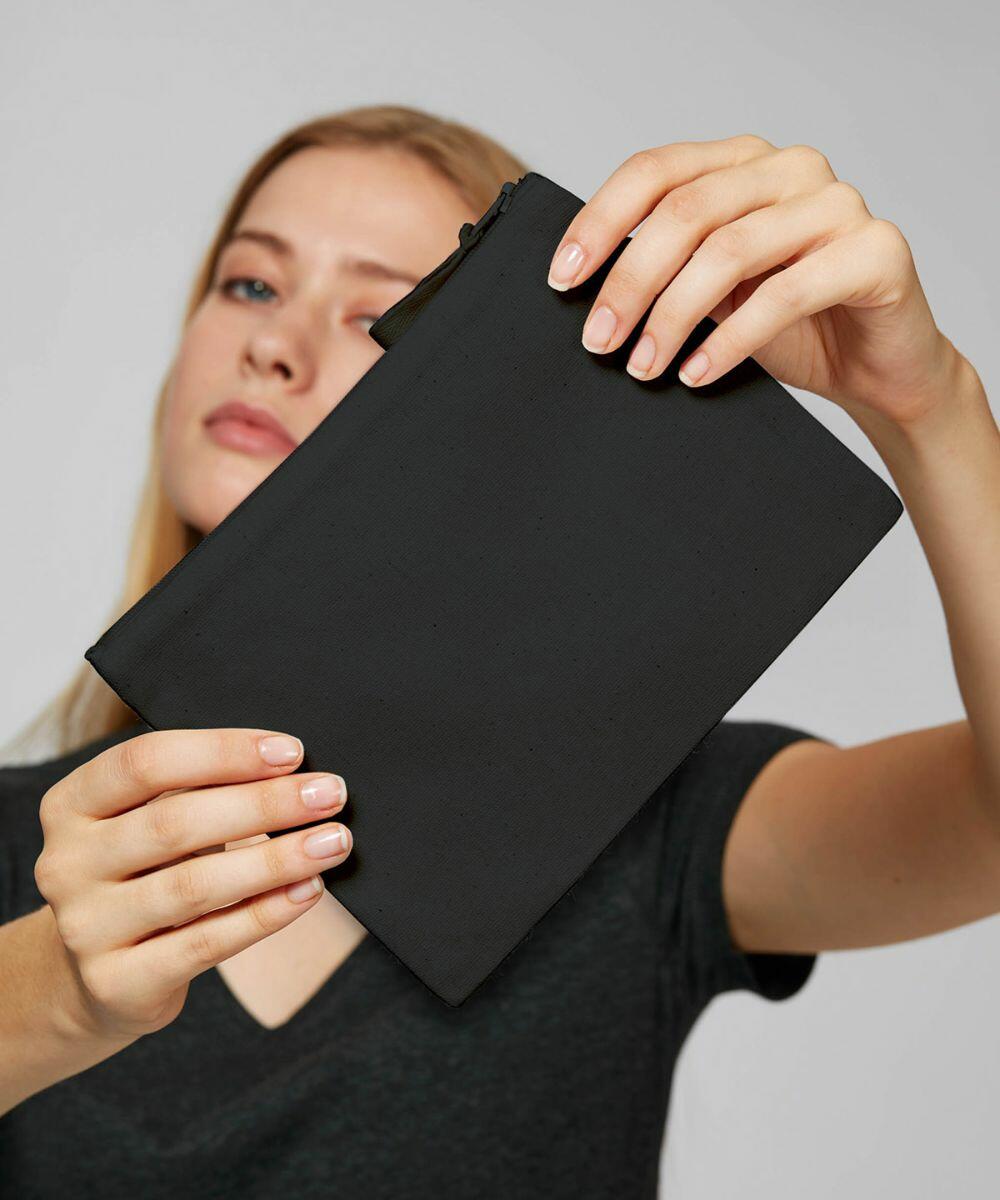 Person holding a Stanley/Stella SX059 black, zippered pouch with both hands, partially covering their face. The background is plain and light-colored.