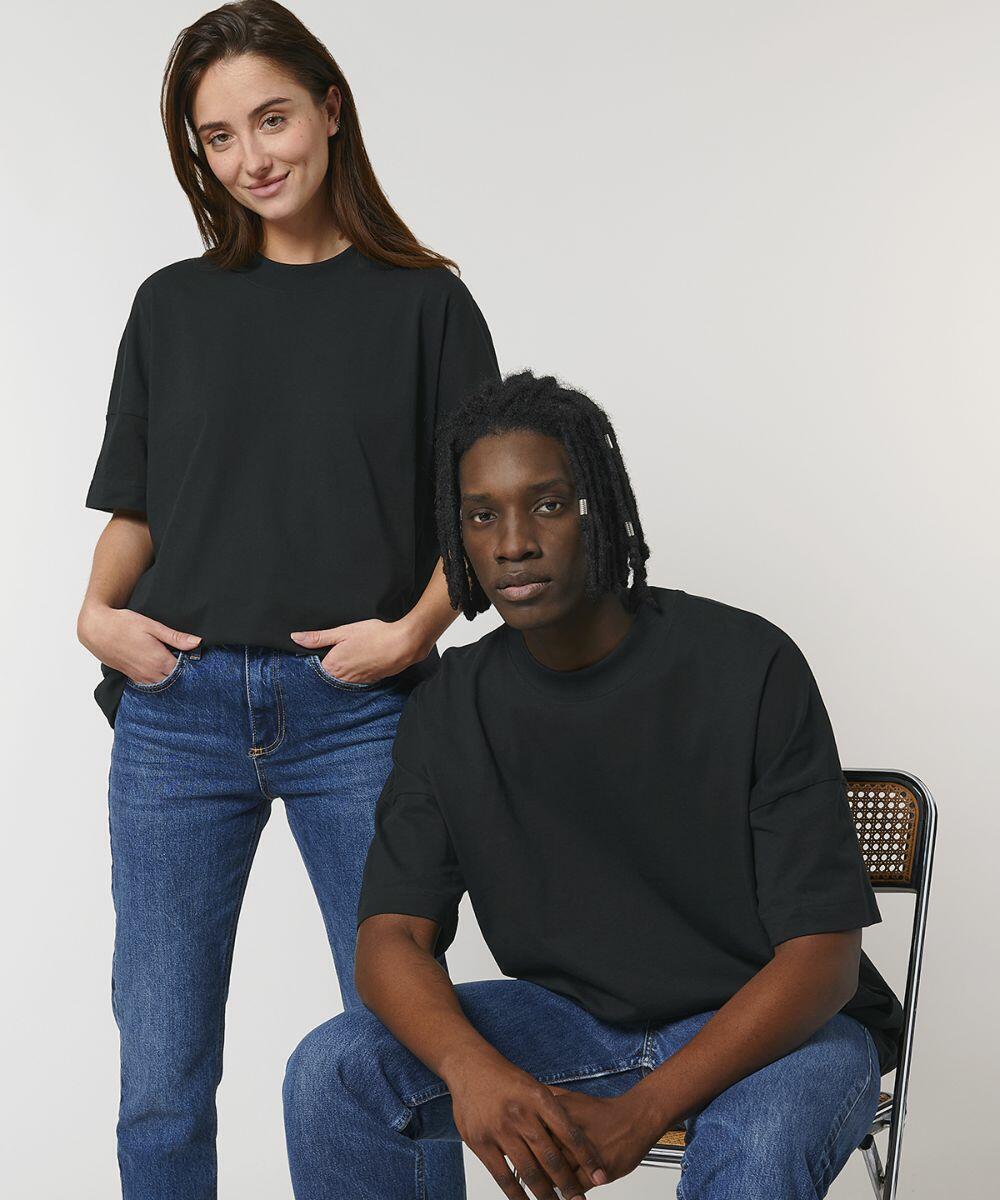 A woman and a man, both wearing Stanley/Stella SX095 black t-shirts and blue jeans, pose for the camera. The woman stands with her hands in her pockets while the man sits on a chair.