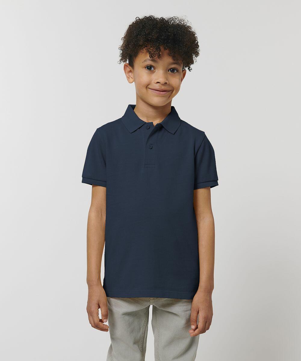 A young boy with curly hair wearing a navy blue Stanley/Stella SX117 polo shirt and light gray pants stands against a plain background, smiling slightly.