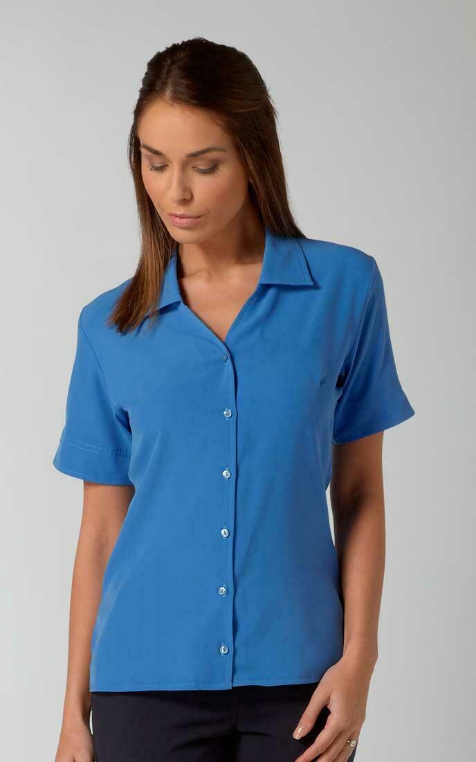 A woman with long brown hair is wearing a buttoned blue short-sleeve Lillie shirt by Vortex and dark pants. She is looking down and has a neutral facial expression, standing against a plain light gray background.