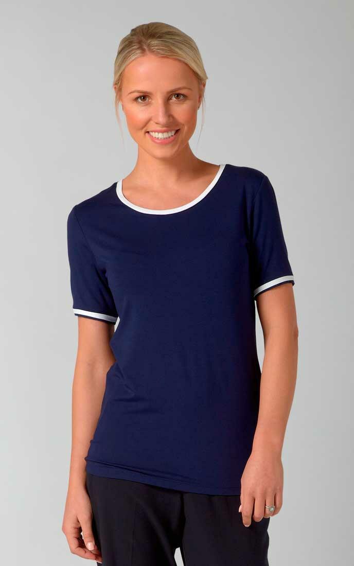 A woman with blonde hair pulled back is smiling at the camera. She is wearing a navy blue short-sleeve Vortex lexie with white trim around the neckline and sleeves. The background is plain and light gray.