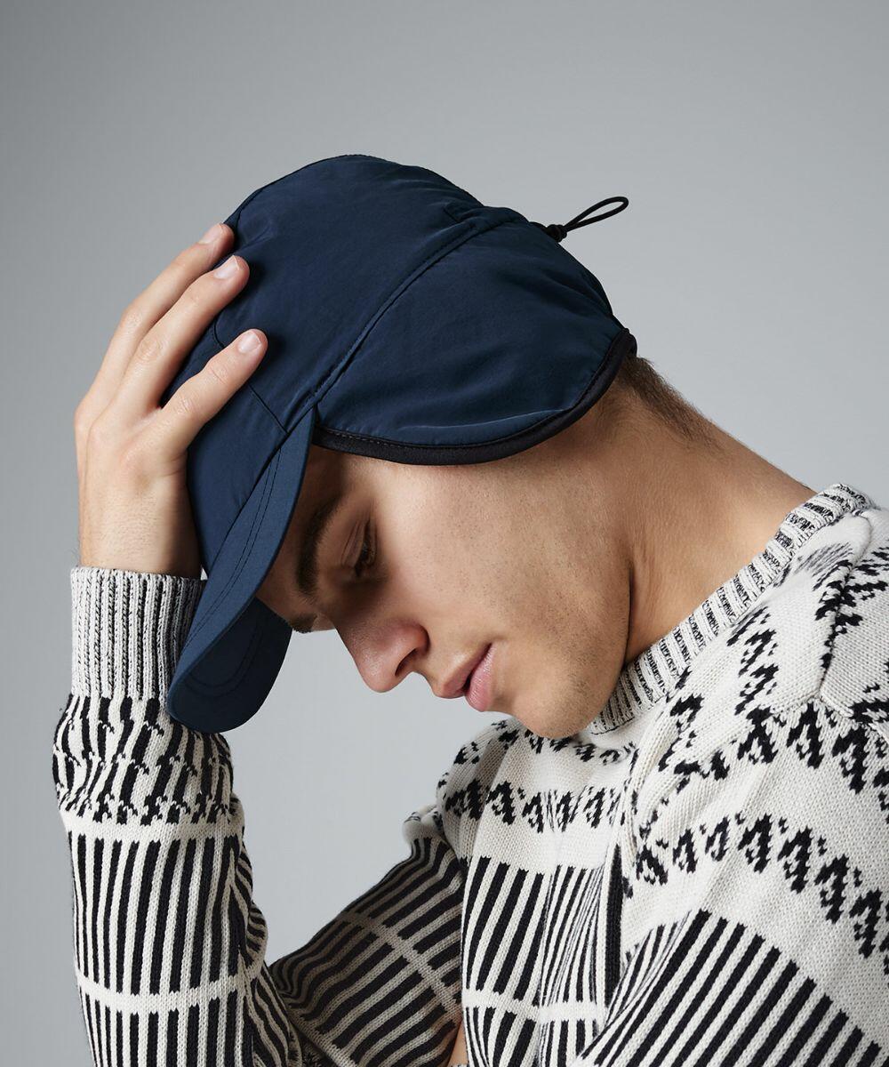 A person wearing a dark blue Beechfield BC355 winter hat with ear flaps and a patterned knit sweater is shown in a side profile. The individual is tilting their head down with one hand resting on the hat, against a light gray background.