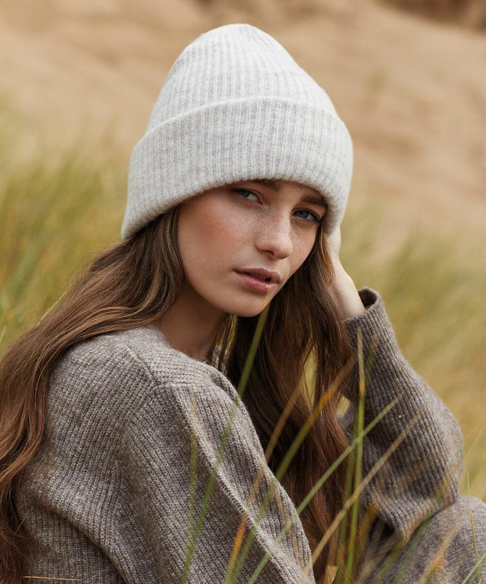 A young woman with long brown hair is sitting outdoors in a grassy area. She is wearing a Beechfield BC386 and a cozy brown sweater. She has a thoughtful expression and is looking directly at the camera while resting her hand against her head.