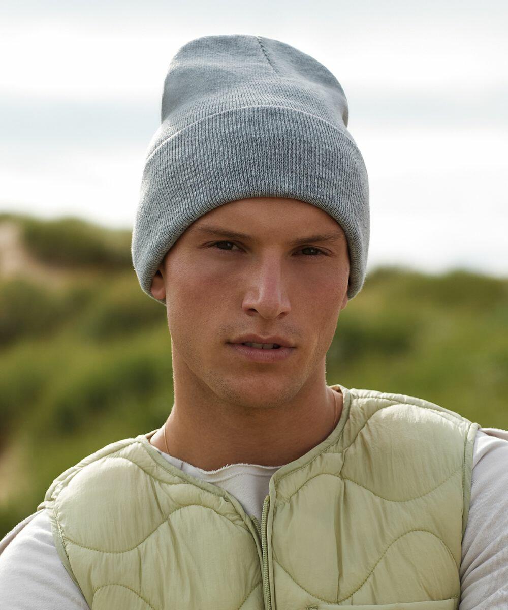 A person wearing a light gray Beechfield BC40R beanie, a beige quilted vest, and a white shirt stands outdoors. The background is blurred with green foliage, suggesting a natural setting.