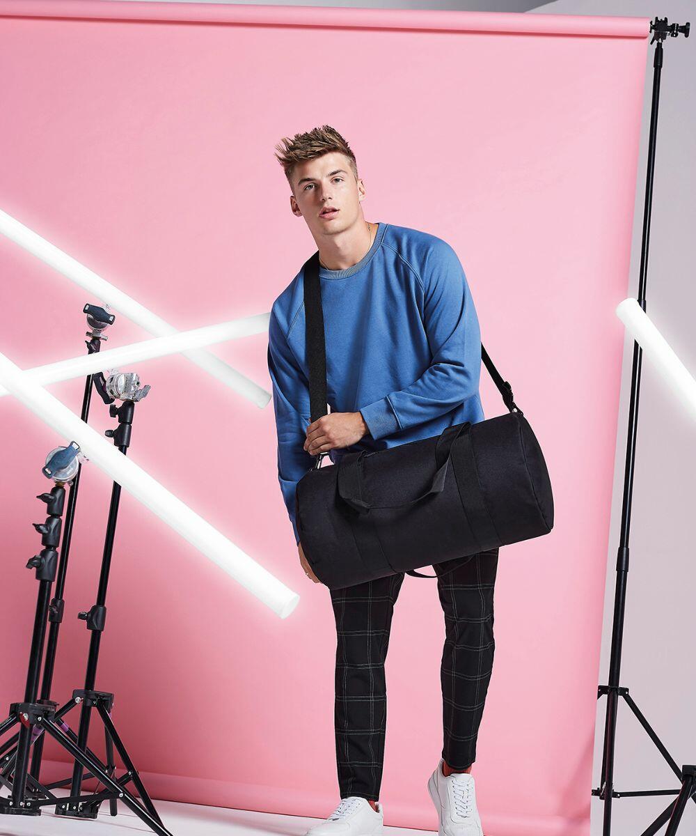 A man is standing against a pink backdrop holding a large black Bagbase B140L over his shoulder. He is wearing a blue long-sleeve shirt, black plaid pants, and white sneakers. Surrounding him are various lighting equipment and fluorescent light tubes.