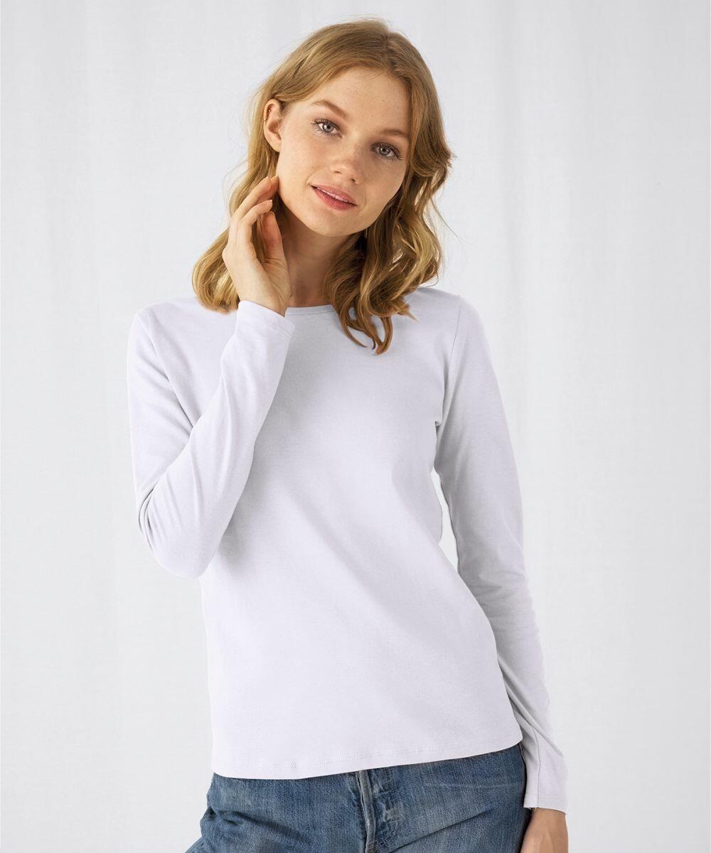 A person with long blonde hair is wearing a plain white B211F long-sleeve shirt from B&C Collection and blue jeans. The individual is posing with one hand touching their neck, against a plain white background. They have a relaxed and friendly expression.