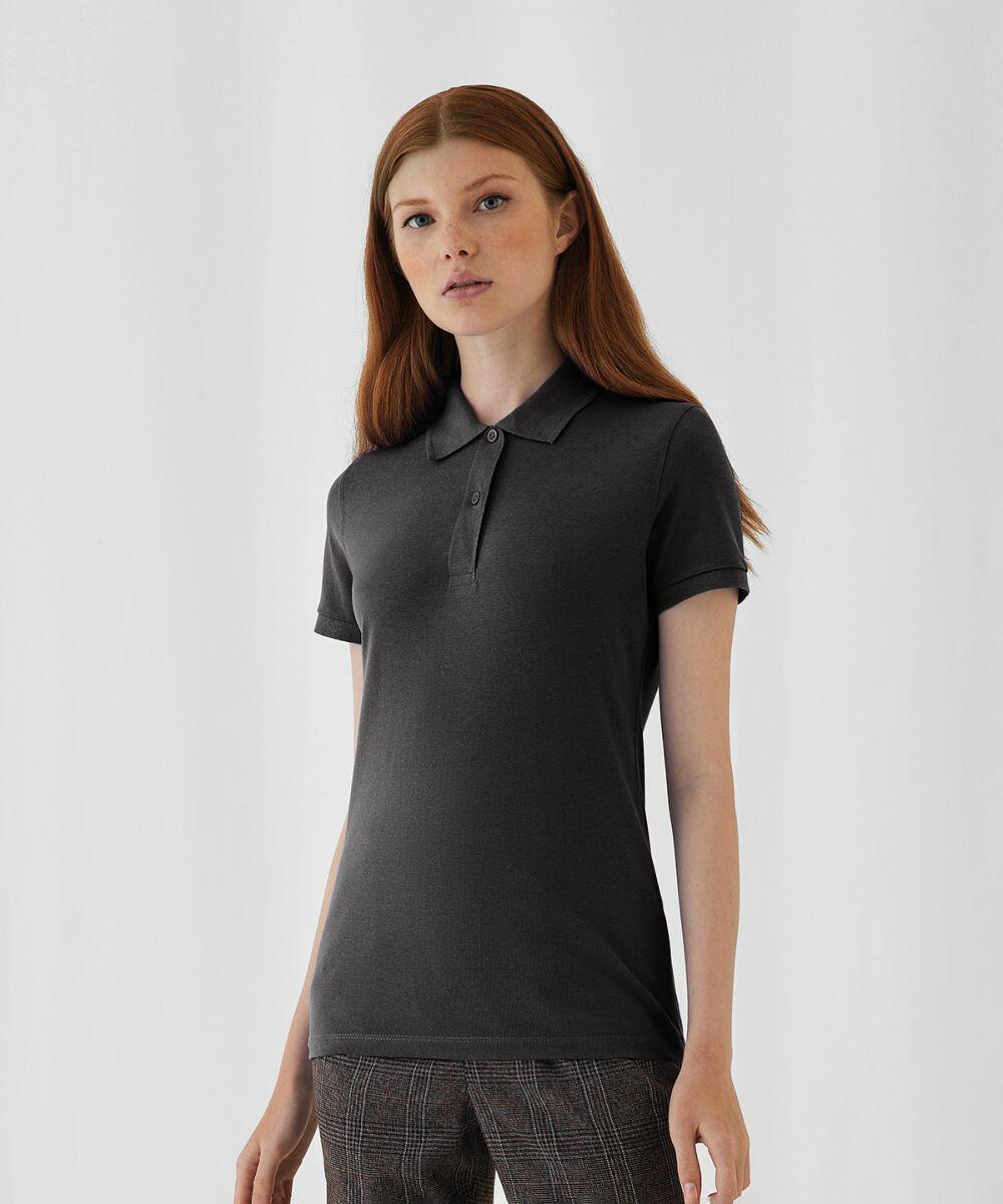 A young woman with long red hair stands against a white background. She is wearing a short-sleeved, charcoal gray B260F by B&C Collection and plaid pants. She looks directly at the camera with a neutral expression.