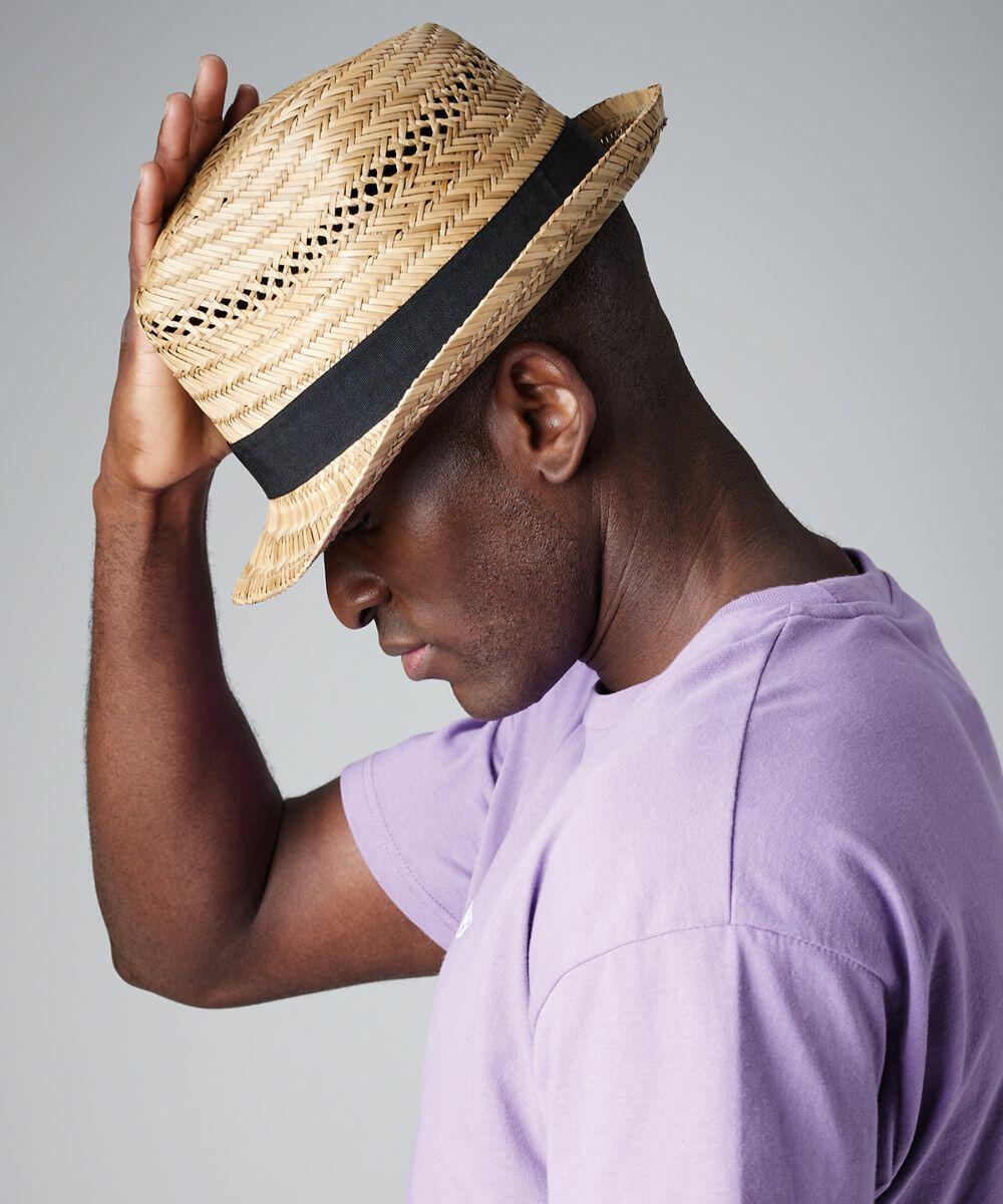 A man wearing a lavender T-shirt and a Beechfield BC730 straw hat with a black band is facing sideways. He is touching the brim of his hat with one hand, tilting his head downward. The background is plain gray.