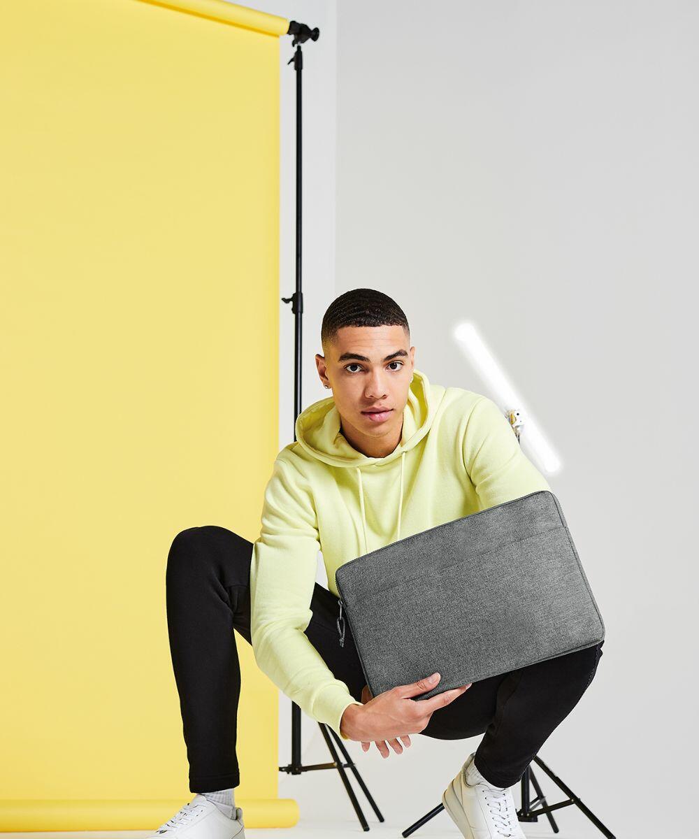 A person in a yellow hoodie and black pants squats in front of a yellow backdrop, holding a gray Bagbase BG068. The individual is wearing white sneakers and is looking at the camera. There is a white strip light in the background.