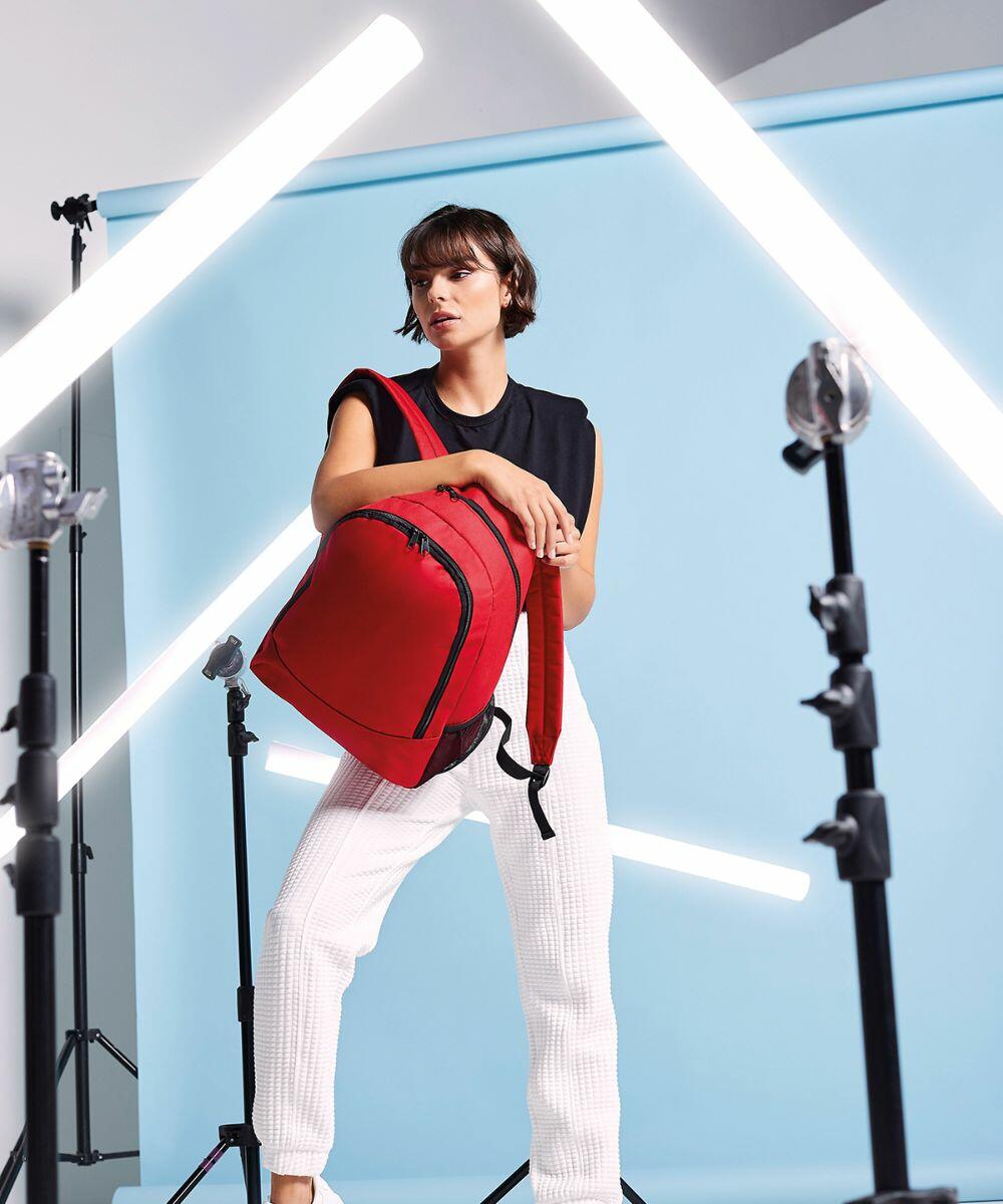 A person stands in a studio setting with a blue backdrop, illuminated by angled LED lights, holding a red Bagbase BG212 and wearing a black top with white pants.
