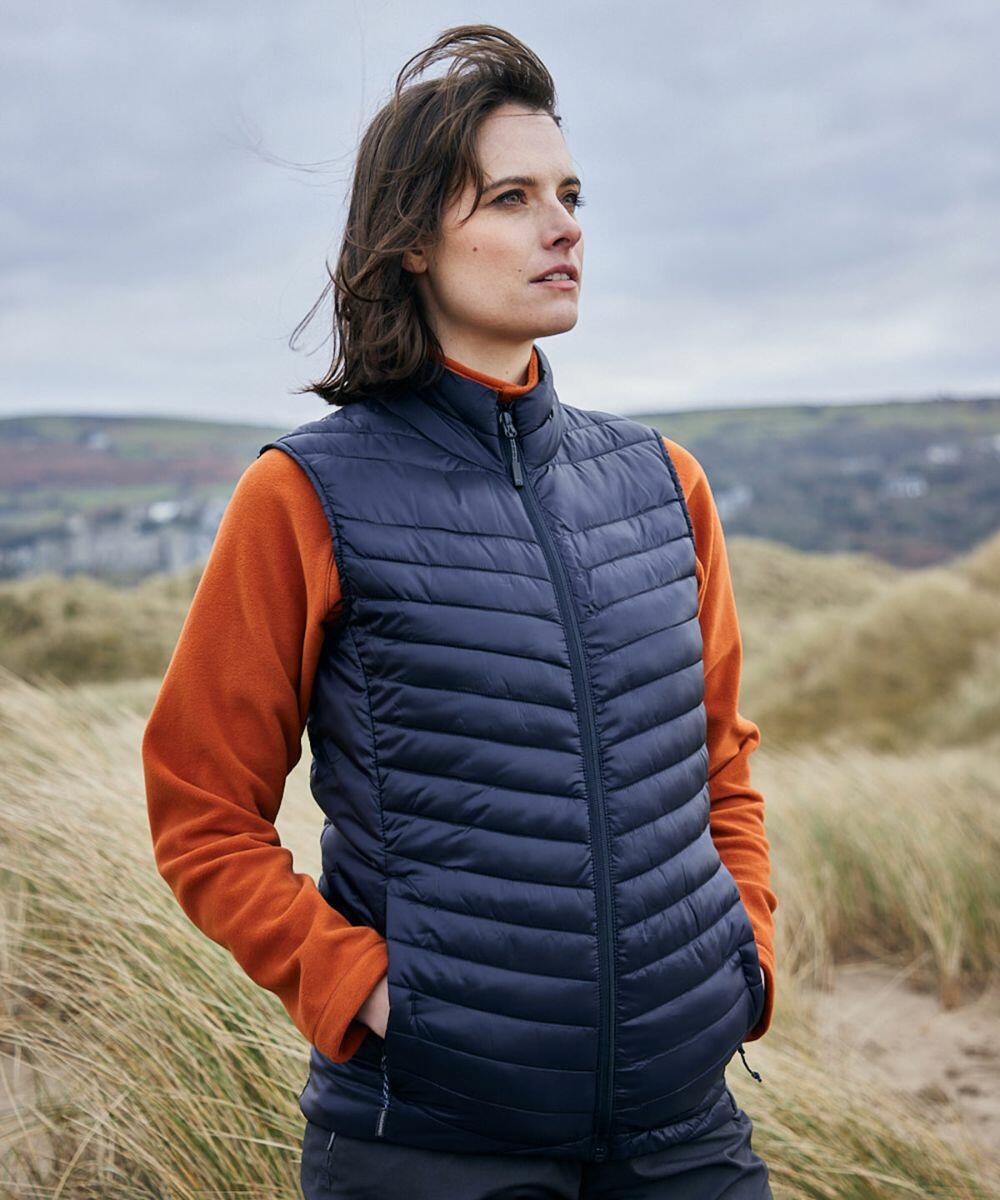 A person wearing an orange long-sleeve shirt and a Craghoppers CR311 puffy navy vest stands outdoors, looking into the distance with a neutral expression. The background features grass and hills under an overcast sky.
