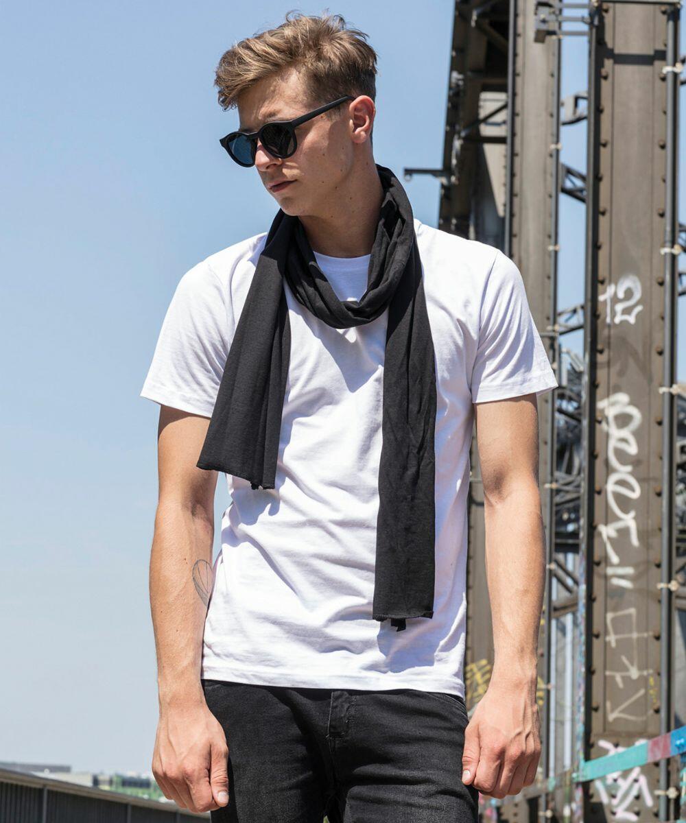 A young man with blond hair wears black sunglasses, a white t-shirt, black scarf, and black pants (BY063 by Build Your Brand), standing outdoors near an industrial structure. The sky is clear with bright sunlight. The structure behind him is metal with some graffiti visible.