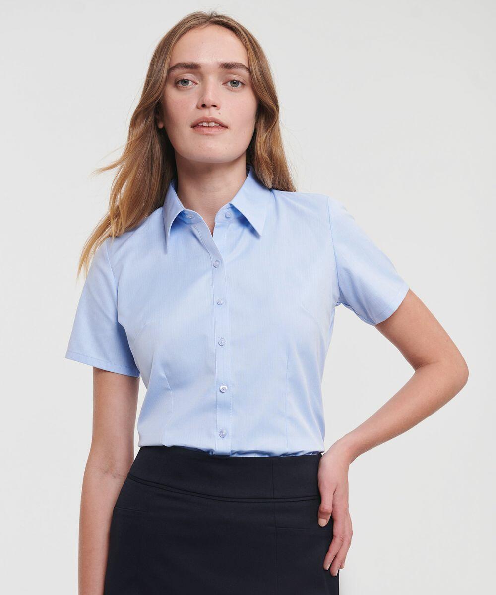 A woman with long hair wearing a light blue J963F button-up shirt from Russell and a dark skirt poses against a plain background.