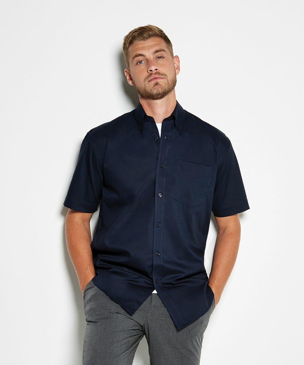 A man with short blond hair and a beard stands against a white background wearing a dark blue KK109 by Kustom Kit short-sleeve button-up shirt and gray pants.