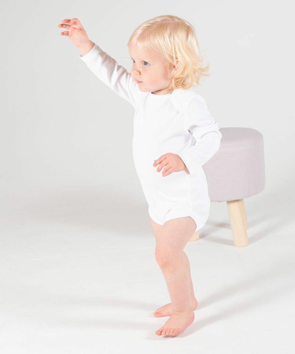 A small child with blond hair wearing a white Larkwood LW52T onesie stands barefoot on a white floor, reaching upward with one hand. A light grey stool is in the background.