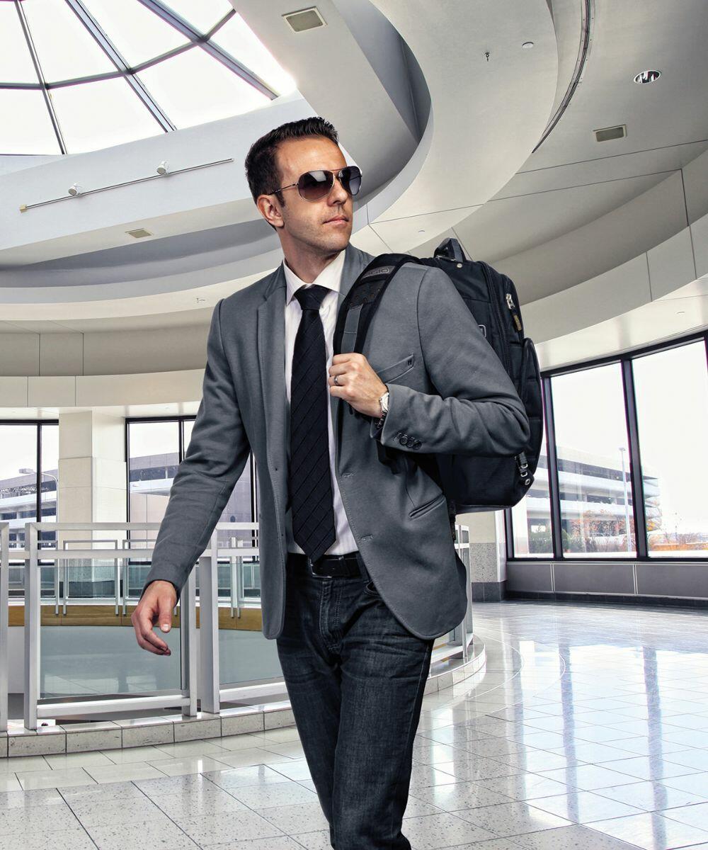 A man in a blazer, tie, and sunglasses walks confidently through a modern airport terminal carrying an Ogio OG027 backpack.