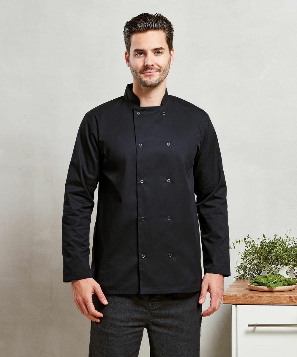 A man in a black double-breasted Premier PR665 chef's jacket stands in front of a light-colored wall, next to a wooden counter holding a green plant.