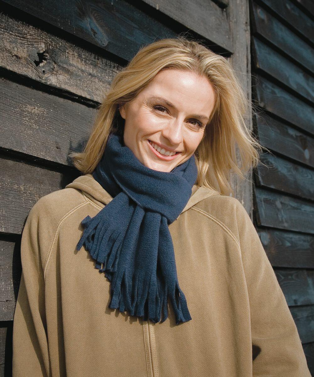 Smiling person with blonde hair wearing a Result R143X tan jacket and blue scarf, standing against a weathered wooden wall.