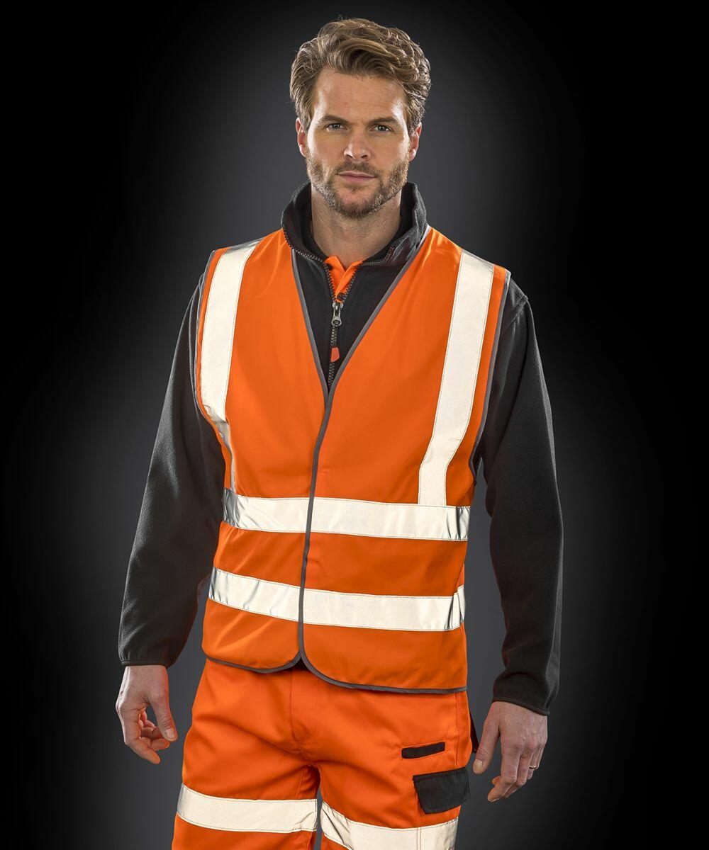 A man wearing a Result R201X orange safety vest with reflective stripes, black long-sleeve shirt, and orange pants, stands against a dark background.