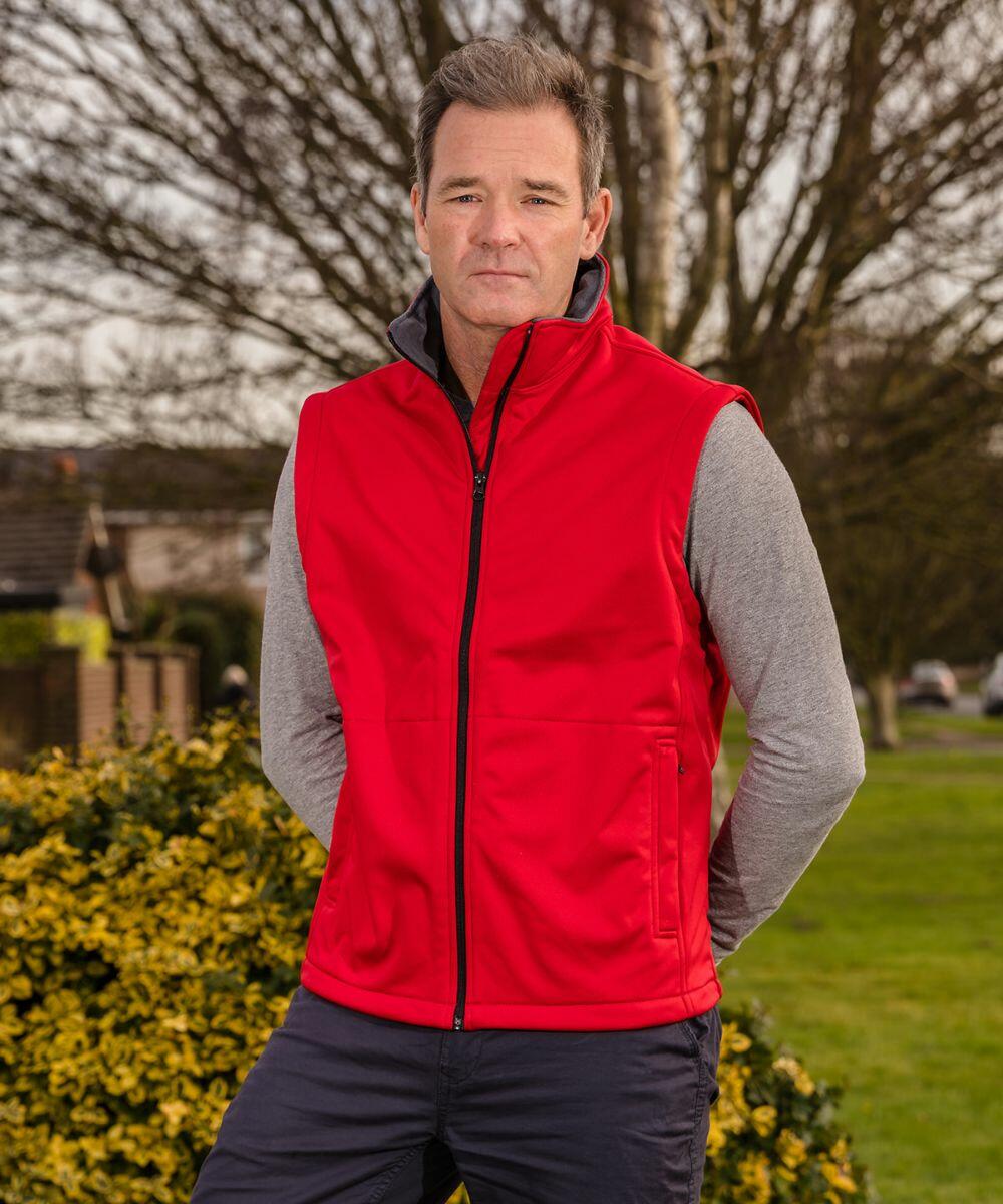 A man stands outdoors wearing a red Result R214X sleeveless jacket over a gray long-sleeve shirt. He is in front of a bush with bare trees and houses visible in the background.
