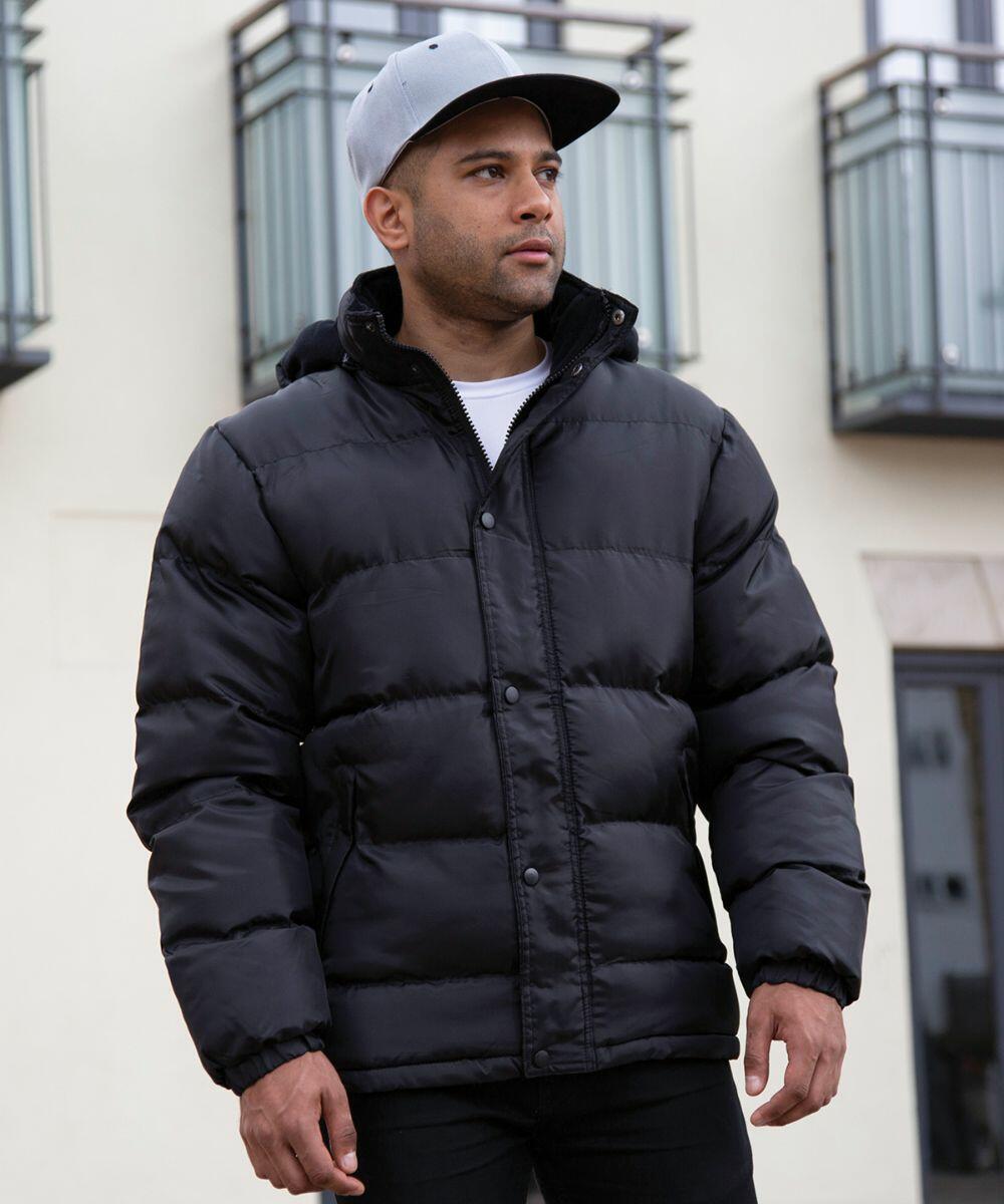 A man wearing a black Result R222X puffer jacket and gray baseball cap stands outside near buildings with balconies.