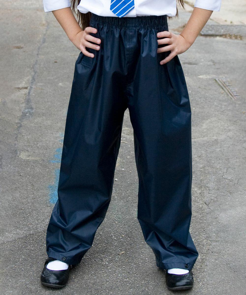 Child in a school uniform stands with hands on hips, wearing a white shirt, blue striped tie, and dark blue Result R226J rain trousers over school shoes. The background is a paved outdoor area.