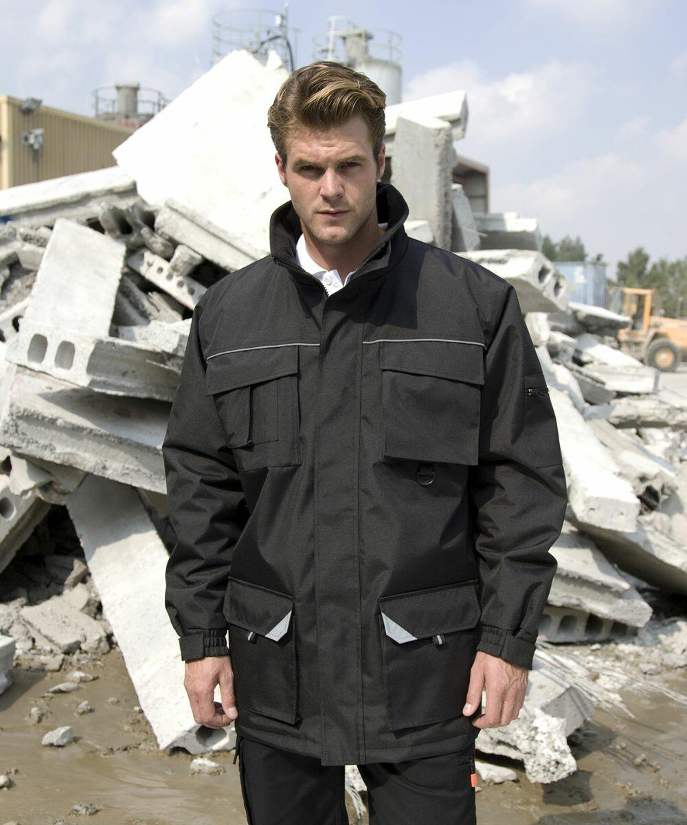 A man wearing a black Result R301X utility jacket stands in front of a pile of concrete debris outdoors.