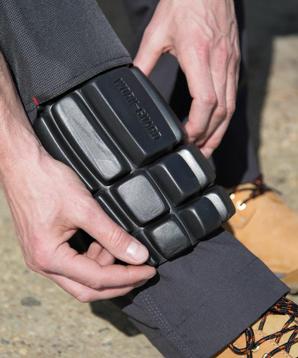 A person adjusts a black Result R322X knee pad over their trousers, wearing brown work boots with orange laces.