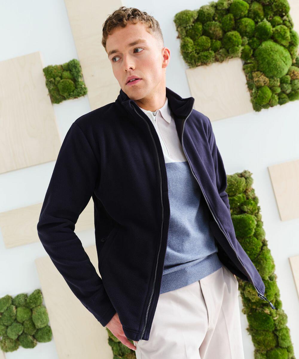 A man wearing a navy **Regatta RG360** jacket, light blue and white polo shirt, and beige pants stands in front of a wall decorated with wooden panels and green moss.