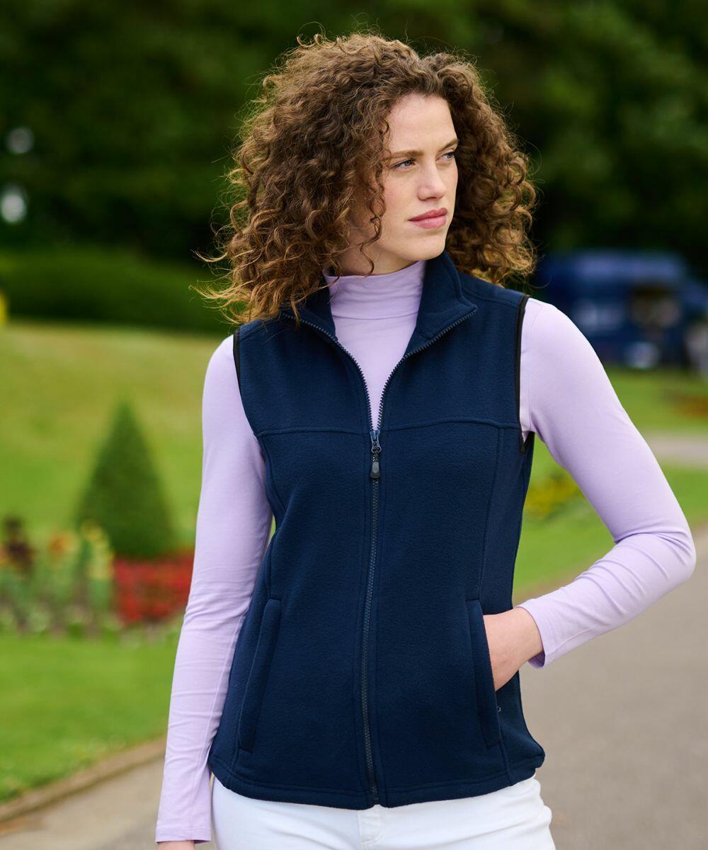 A person with curly hair is wearing a navy blue Regatta RG184 zip-up vest over a light purple long-sleeve shirt, standing outdoors in a garden setting.