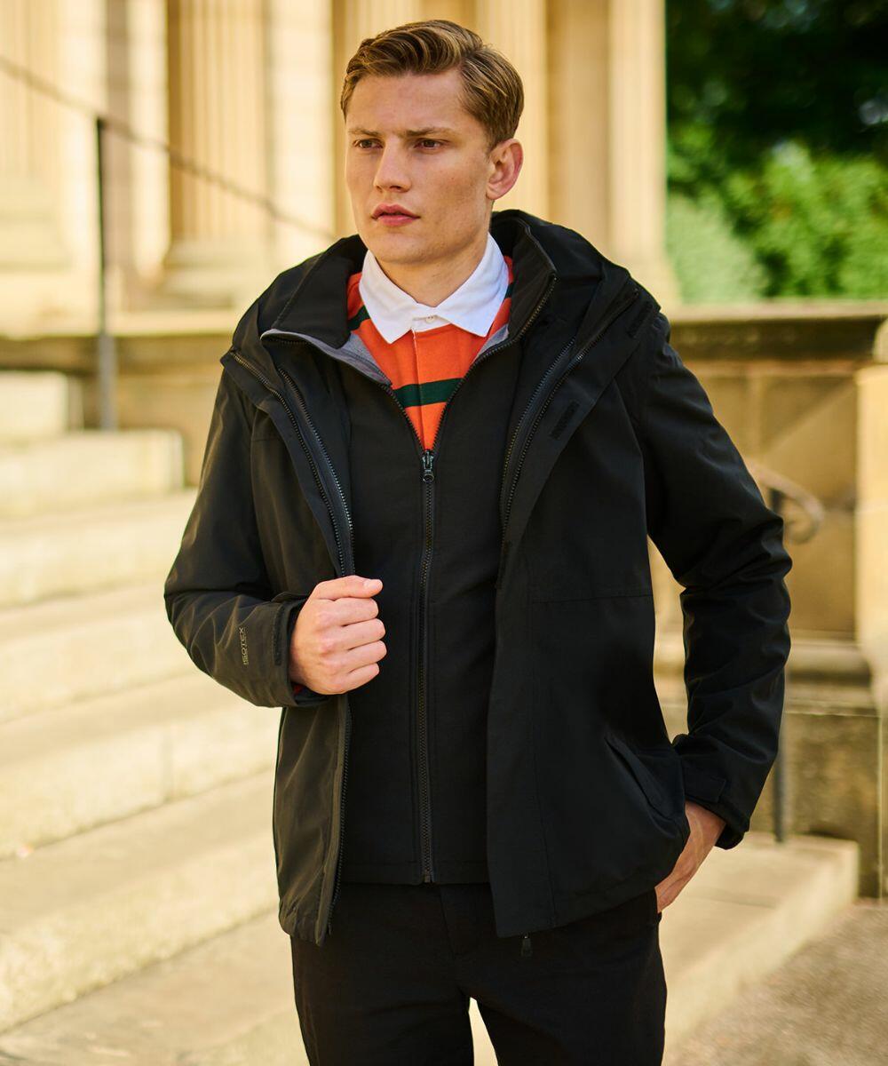 A man stands outdoors wearing a black RG191 jacket by Regatta with a striped collar, looking to his left. Steps and columns of a building are visible in the background.