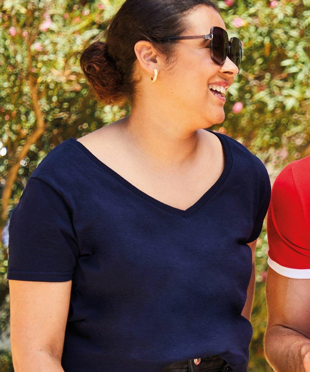 A woman wearing Fruit of the Loom's SS047 navy blue V-neck t-shirt smiles and looks to her right. She is standing outdoors with greenery in the background.