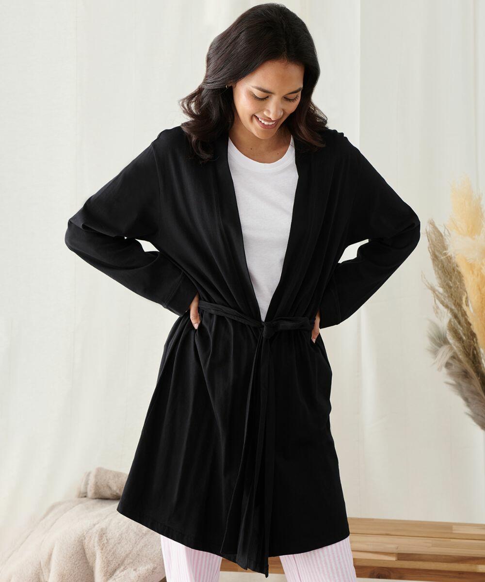 A woman stands indoors wearing a black robe over a white shirt and pink striped pants, smiling and looking down with her hands on her hips. Neutral background with a beige blanket and decorative grasses. She is feeling cozy in the TC050 from Towel City.